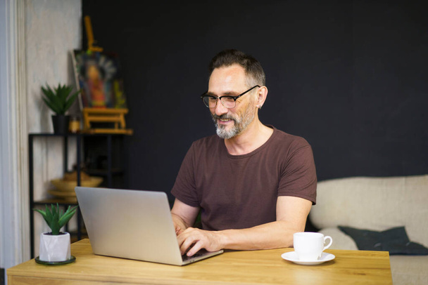 mature man in glasses sitting at his modern desk at own living room with dark wall and typing on laptop - Zdjęcie, obraz