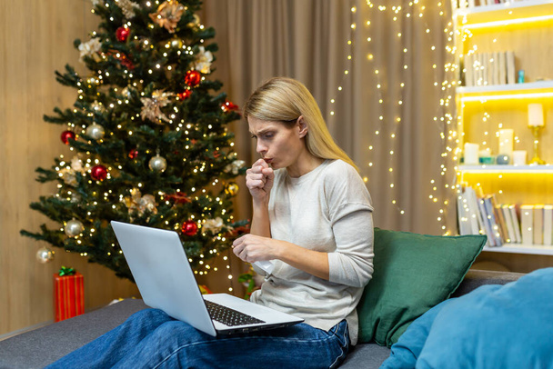Woman sick on Christmas working with laptop on New Year holidays coughing sitting on sofa at home in living room near Christmas tree. - 写真・画像