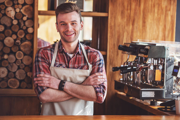 Bello giovane barista in grembiule sta guardando la fotocamera e sorridente mentre in piedi con le braccia incrociate al bancone del bar al caffè
 - Foto, immagini