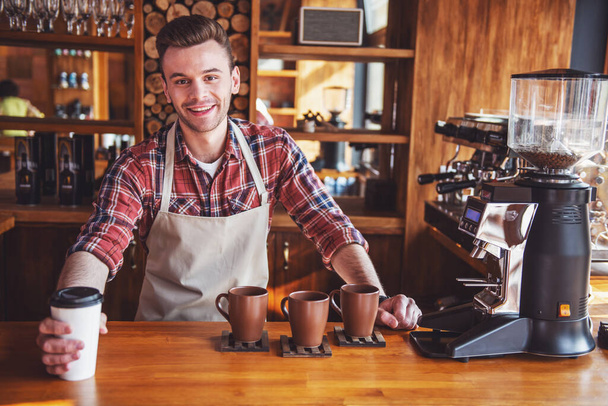 Csinos fiatal barista kötény van giving egy csésze kávét, látszó-on fényképezőgép, és mosolyogva-állva a bárban számláló kávézóban - Fotó, kép