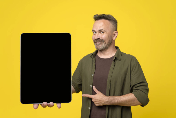 Happy smiled stubble and mustached mature man in green shirt pointing to his tablet pc with black mock-up screen holding in hand on yellow background - Fotografie, Obrázek