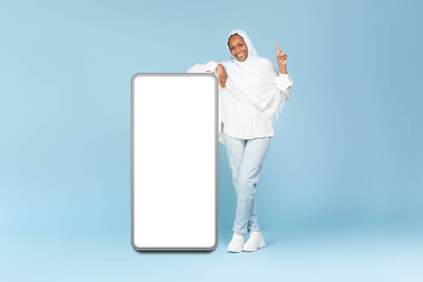 Мобильное приложение, отличное предложение. Полная длина тела черной мусульманки, указывающей пальцем вверх, опираясь на большой смартфон с пустым экраном, стоящий на синем фоне студии. Mockup - Фото, изображение
