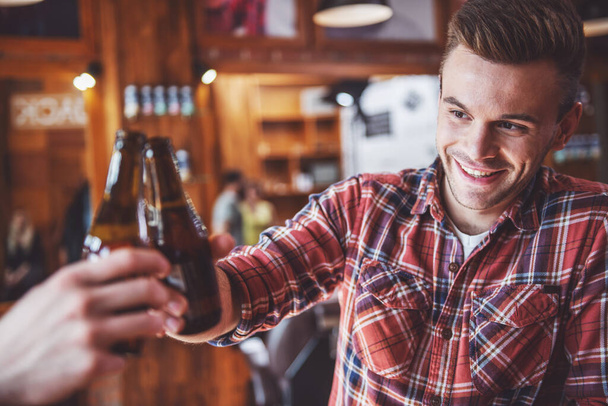 Jovem bonito está batendo garrafa de cerveja e sorrindo, sentado em um café urbano moderno
 - Foto, Imagem