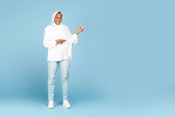 Special offer. Full length shot of black muslim lady pointing fingers aside at copy space standing over blue studio background. Positive islamic woman showing empty mockup place for ad, banner - 写真・画像