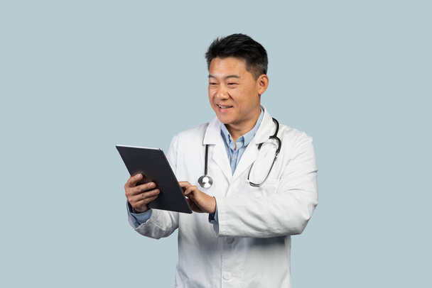 Χαρούμενος μεσήλικας Κινέζος θεραπευτής με λευκό παλτό δακτυλογραφώντας σε tablet, απομονωμένος σε μπλε φόντο, στούντιο, χώρος αντιγραφής. Νέα εφαρμογή για τη θεραπεία, ιστοσελίδα για την υγειονομική περίθαλψη, φάρμακα και νέα φυσιολογικά - Φωτογραφία, εικόνα