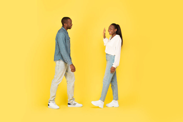 Cheerful Black Couple Walking Meeting Each Other, Woman Waving Hand Posing Over Yellow Studio Background. Friendship, Relationship And Communication. Hello Concept. Full Length Shot - Photo, Image