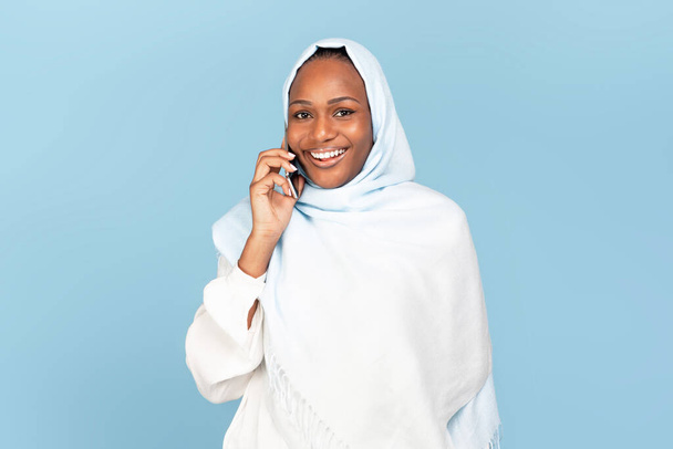 Iloinen afrikkalainen amerikkalainen nainen hijabissa soittaa matkapuhelimella, puhuu kollegan tai ystävän kanssa älypuhelimella sinisellä taustalla ja hymyilee kameralle - Valokuva, kuva