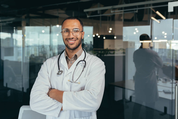 Portrait of a young handsome male hispanic doctor. Standing in the glass hall of the hospital wearing glasses, a white coat and a stethoscope. He looks at the camera, crosses his arms, smiles. - Foto, imagen