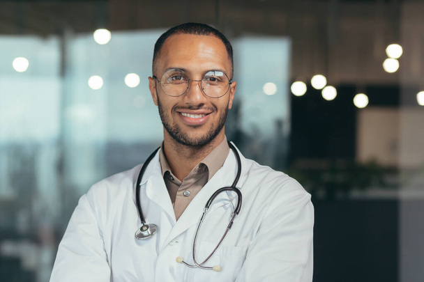 Fotografía de cerca. Retrato de un joven guapo médico hispano. De pie en la sala de cristal del hospital con gafas, una bata blanca y un estetoscopio. Mira a la cámara, sonríe - Foto, Imagen