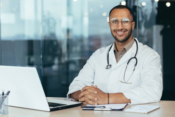Portrait of young hispanic male pediatrician doctor. Sitting in the office at a table with documents and a laptop, wearing glasses and a stethoscope in a white coat, looking at the camera, smiling. - Foto, Imagen