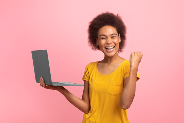 Iloinen innoissaan nuori sekarotuinen nainen kihara tukka iloitsee verkossa menestystä kannettavan, jolloin voitto ele, eristetty vaaleanpunainen tausta, studio. Hyviä uutisia, tunteita, valtava myynti, mainos ja tarjous - Valokuva, kuva