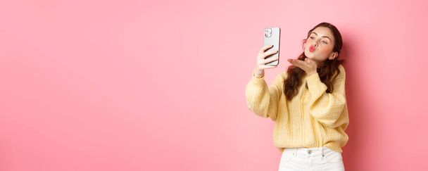 Beautiful flirty girl taking selfie on smartphone, sending air kiss during video chat, standing against pink background. Copy space - Foto, Imagen
