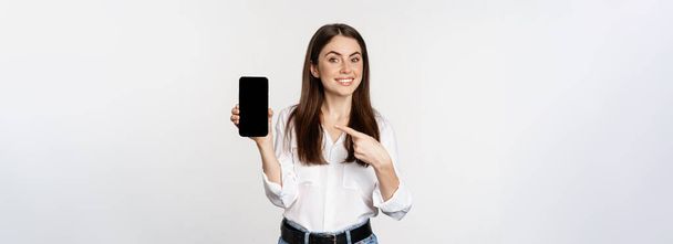 Glückliche schöne Frau lächelt, zeigt auf Handy-Bildschirm, zeigt Werbung, Website, steht über weißem Hintergrund. - Foto, Bild