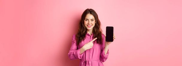 Young caucasian woman in stylish dress, pointing finger at smartphone screen and smiling, showing promo deal online, pink background. - Photo, Image