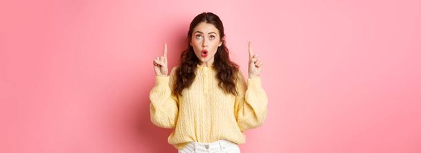 Surprised caucasian girl gasping, saying wow and looking with interest on top, pointing fingers up, standing in casual clothes, pink background. - Photo, Image