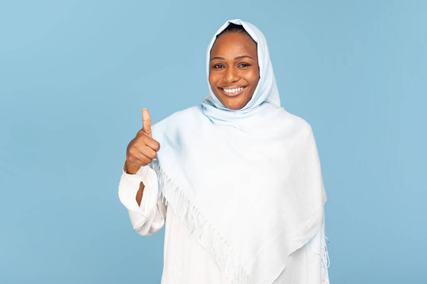 Gelukkige Afrikaanse amerikaanse moslim vrouw die duim omhoog op blauwe studio achtergrond, kijkend en glimlachend op camera. Vrolijke zwarte dame in hijab aanbevelen product, service of aankoop - Foto, afbeelding
