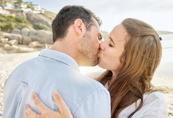 Happy, kiss and couple on a beach with love from engagement announcement or anniversary. Happiness, relax and care of a woman and man together by the sea and ocean outdoor with commitment. - Zdjęcie, obraz
