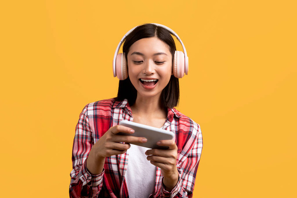 Διασκέδαση και online παιχνίδια. Χαρούμενη Ασιάτισσα κυρία με μοντέρνα ασύρματα ακουστικά που παίζει στο smartphone, στέκεται πάνω από το κίτρινο φόντο του στούντιο - Φωτογραφία, εικόνα