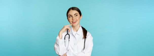 Junge Ärztin, Krankenhausangestellte im weißen Kittel, nachdenklich und wegschauend, Lösung suchend, vor toquoisem Hintergrund stehend. - Foto, Bild