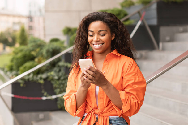 Veselý tisíciletý africký americký žena v příležitostném chatování na smartphonu, čtení zprávy, chat a blog v City Street. Nová mobilní aplikace, sociální sítě a komunikace vzdáleně se zařízením - Fotografie, Obrázek