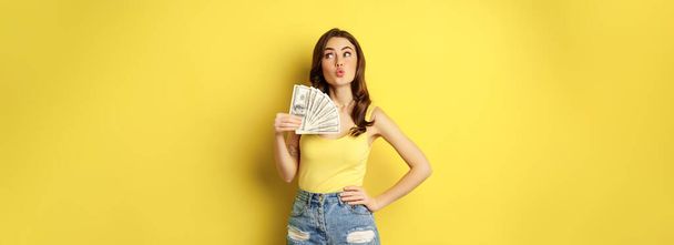 Shopping, credit and money concept. Young brunette woman showing cash and smiling pleased, standing over yellow background. - 写真・画像