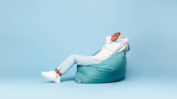 Lunghezza completa della donna musulmana nera rilassata seduta sulla poltrona beanbag, che si tiene per mano dietro la testa, avendo rotto sullo sfondo blu, design banner, panorama - Foto, immagini