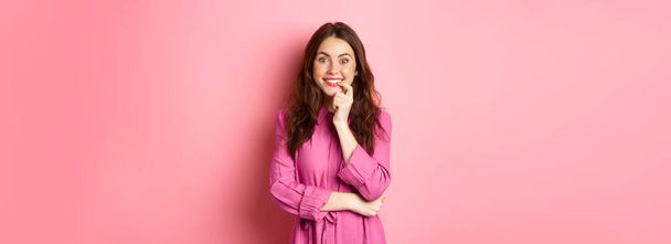 Beautiful excited girl smiling, biting finger and looking with tempted face at camera, having interesting idea, looking at something intriguing, standing against pink background. - Photo, Image