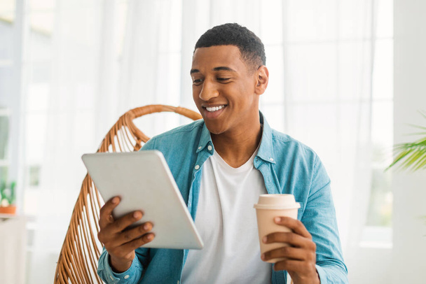 Lachende jonge zwarte man in casual chatten op tablet, Bekijk video, genieten van afhaaldrankje, zitten in een fauteuil in lichte kamer interieur. Rust in de vrije tijd, koffiepauze met nieuwe app, surfen op internet - Foto, afbeelding