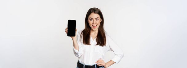 Mujer sonriente feliz mostrando interfaz de aplicación de teléfono inteligente, pantalla del teléfono móvil, demostrando oferta promocional, de pie sobre fondo blanco. - Foto, imagen