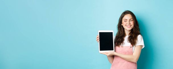 Image of happy young girl showing digital tablet screen and smiling proud, showing your logo on display, standing over blue background. - Photo, Image