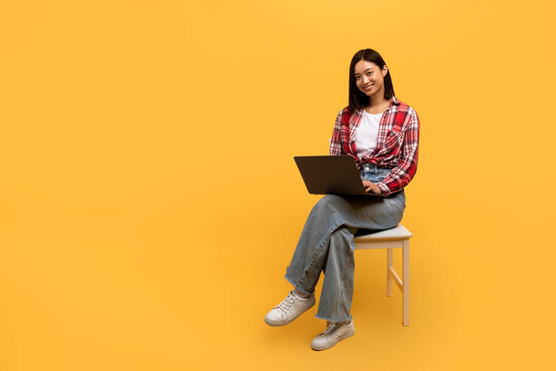 Positieve Koreaanse dame student met behulp van laptop, surfen op internet tijdens het zitten op stoel over gele studio achtergrond, kopiëren ruimte. E-learning, online lezingen en cursussen - Foto, afbeelding