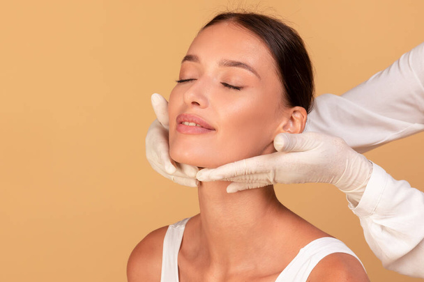 Young lady with smooth skin receiving inspecting of face in medical centre, cosmetologist in white rubber gloves touching neck, chin, examining over beige background - Valokuva, kuva
