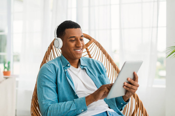 Cieszę się szczęśliwy młody czarny mężczyzna w słuchawkach casual i bezprzewodowych czatujących w sieciach społecznościowych na tablecie, oglądać filmy siedzi na krześle w jasnym wnętrzu pokoju. Blog, nowa aplikacja, odpoczynek w domu w weekend - Zdjęcie, obraz
