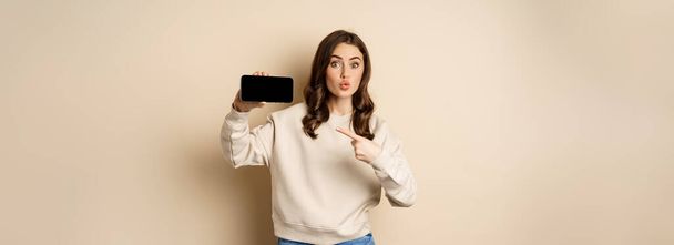 Wizerunek dziewczyny wskazującej palcem, pokazującej ekran w poziomie telefonu komórkowego i wyglądającej na zdumioną aplikacją, stojącej nad beżowym tłem. - Zdjęcie, obraz
