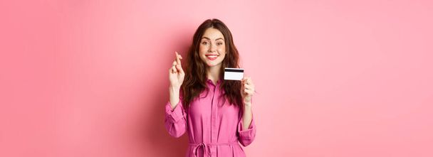 Εικόνα της ελπιδοφόρας νεαρής γυναίκας που δείχνει πλαστική πιστωτική κάρτα και σταυρώνουν τα δάχτυλα για καλή τύχη, κάνοντας ευχές και χαμογελώντας, στέκεται πάνω σε ροζ φόντο. - Φωτογραφία, εικόνα