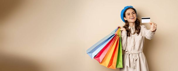 Portrat of trendy feminine girl posing with shopping bags from store and credit card, paying contactless, buying with discount on sale, beige background. - Φωτογραφία, εικόνα