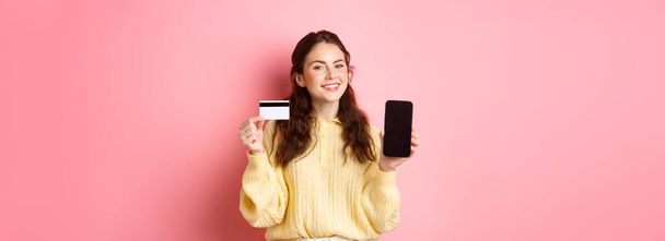 Technologie und Online-Shopping. Porträt der schönen Dame zeigt Handy-Bildschirm und Plastikkreditkarte, lächelt zufrieden, empfehle App, steht über rosa Hintergrund. - Foto, Bild