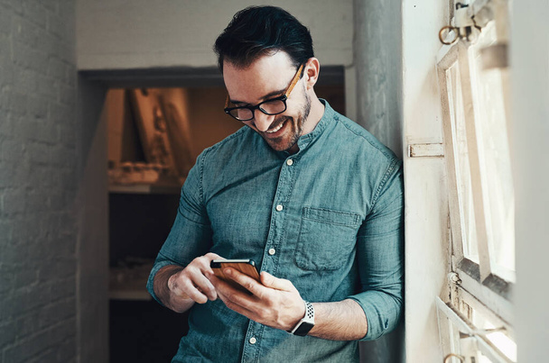 Nie dostałbym dziś lepszego SMS-a. przystojny młody biznesmen uśmiechnięty podczas korzystania ze smartfona w nowoczesnym biurze - Zdjęcie, obraz