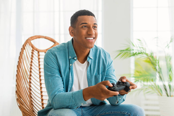 Cieszę się szczęśliwy młody czarny mężczyzna w dżinsach z joystick gra online, baw się, cieszyć gadżet, siedzi na krześle w świetle wnętrza pokoju. Nowa aplikacja do rozrywki w domu sam podczas epidemii covid-19 - Zdjęcie, obraz