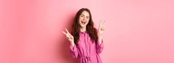 Positief mooi meisje in stijlvolle jurk, tonen v-teken vrede en glimlachen blij, poseren in de buurt van roze achtergrond. - Foto, afbeelding