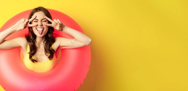 Portret van aantrekkelijk modern meisje in trendy roze strand zwemmen ring, tonen vrede v-teken en glimlachen blij, staande over gele achtergrond. - Foto, afbeelding