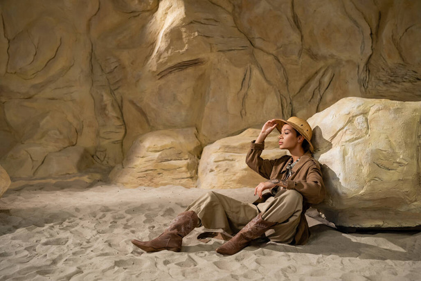 teljes hossza archeológus beállítása szafari kalap, miközben ül a homok közelében kő - Fotó, kép