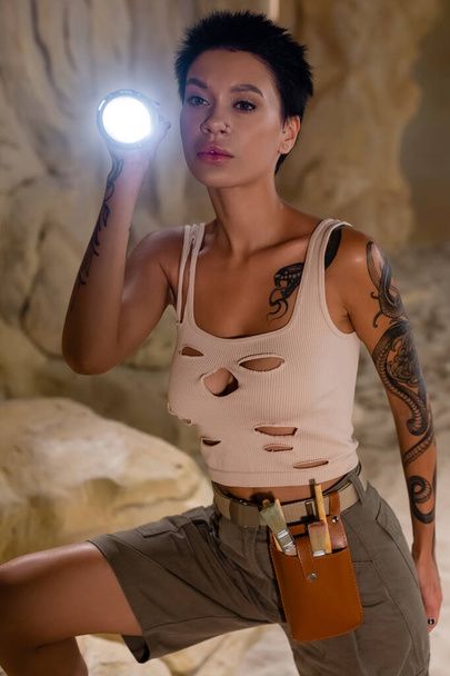 sexy archeologo bruna in canotta in piedi con torcia elettrica nella grotta del deserto - Foto, immagini