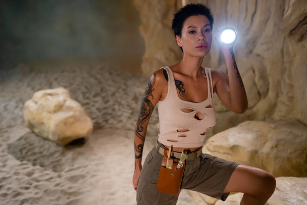 sexy archaeologist in tank top and waist bag with brushes holding flashlight in cave - Photo, Image