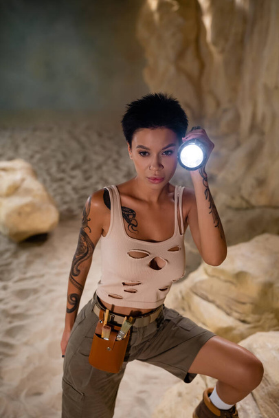 tattooed brunette archaeologist with waist bag holding flashlight in cave - Photo, Image