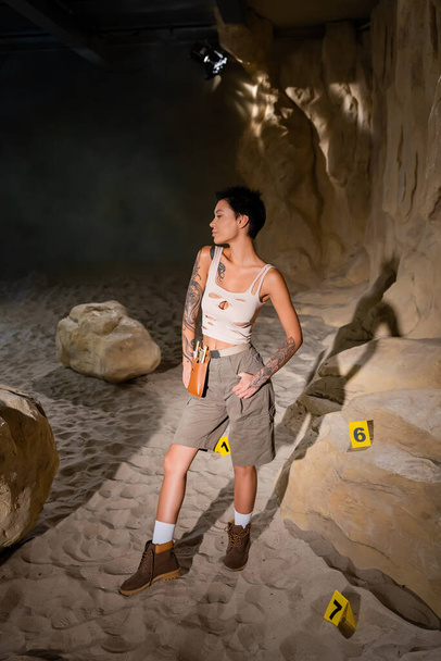 täyspitkä seksikäs tatuoitu arkeologi seisoo luolassa lähellä numeroituja kortteja hiekassa - Valokuva, kuva