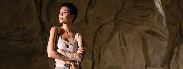 tatuoitu arkeologi seksikäs toppi seisoo ristissä kädet ja katselee pois luolassa, banneri - Valokuva, kuva