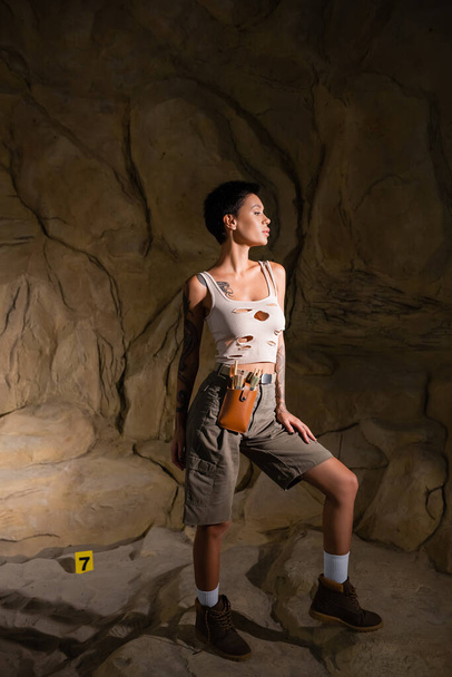 täyspitkä tatuoitu brunette arkeologi toppi ja shortsit etsivät pois pimeässä luolassa - Valokuva, kuva