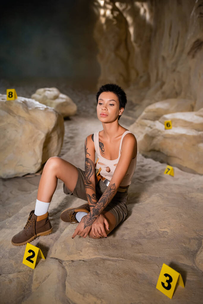full length of sexy and thoughtful archaeologist sitting near numbered marks in cave - Photo, Image