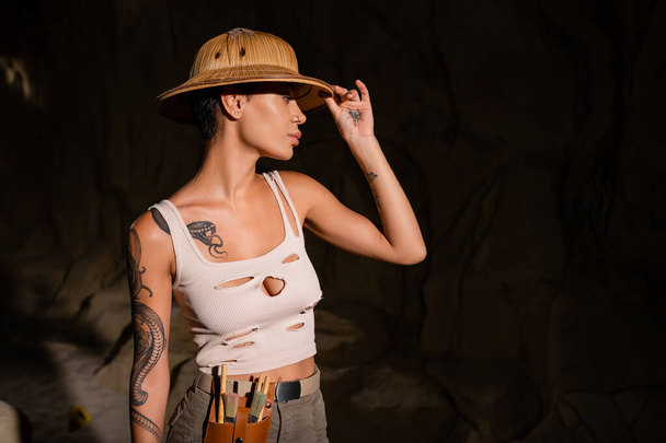 tattooed archaeologist with tools in waist bag adjusting safari hat while looking away in dark desert - Фото, изображение